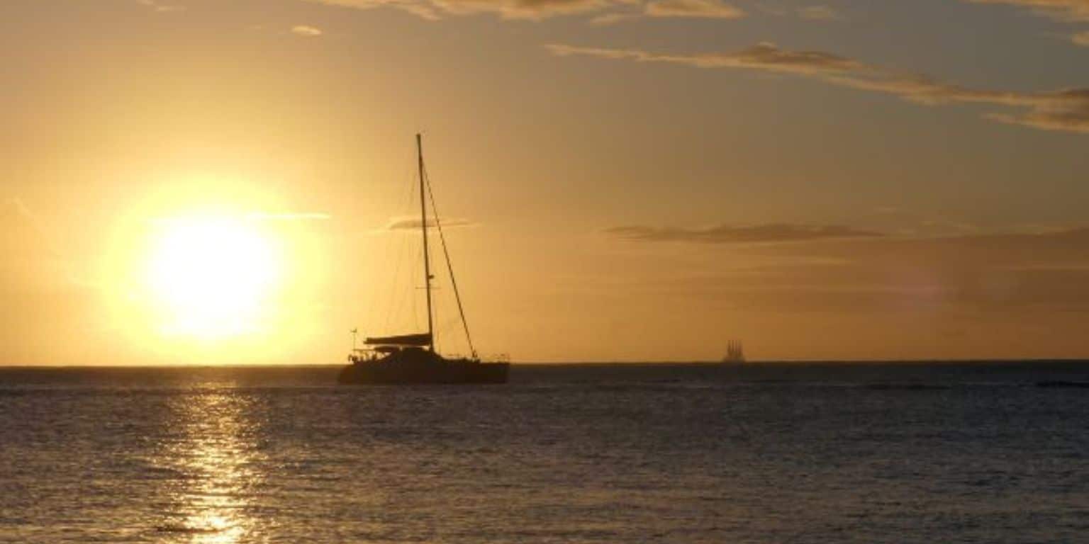 Voilier naviguant au coucher du soleil sur les eaux paisibles de Tahiti, avec une lumière dorée reflétant sur l’océan. Une scène évocatrice de voyage, d’évasion et de la beauté naturelle de la Polynésie française, berceau des perles de Tahiti signées Poe Ora Création.